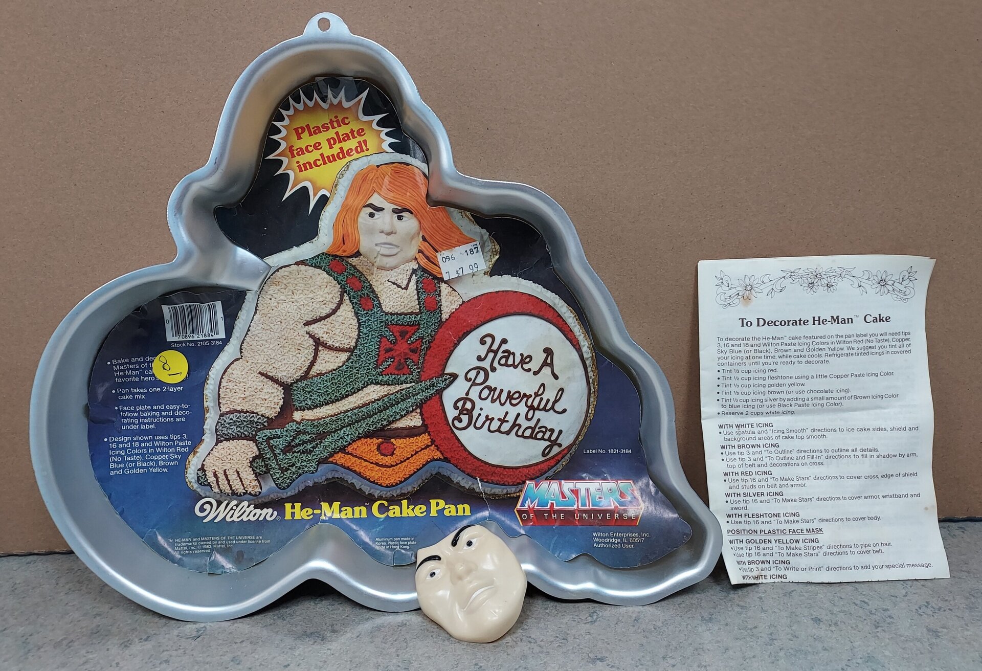 A He-Man-shaped cake pan with packaging showing a decorated He-Man cake and the text “Have A Powerful Birthday.” Included are a plastic He-Man face plate and a decorating instruction sheet.