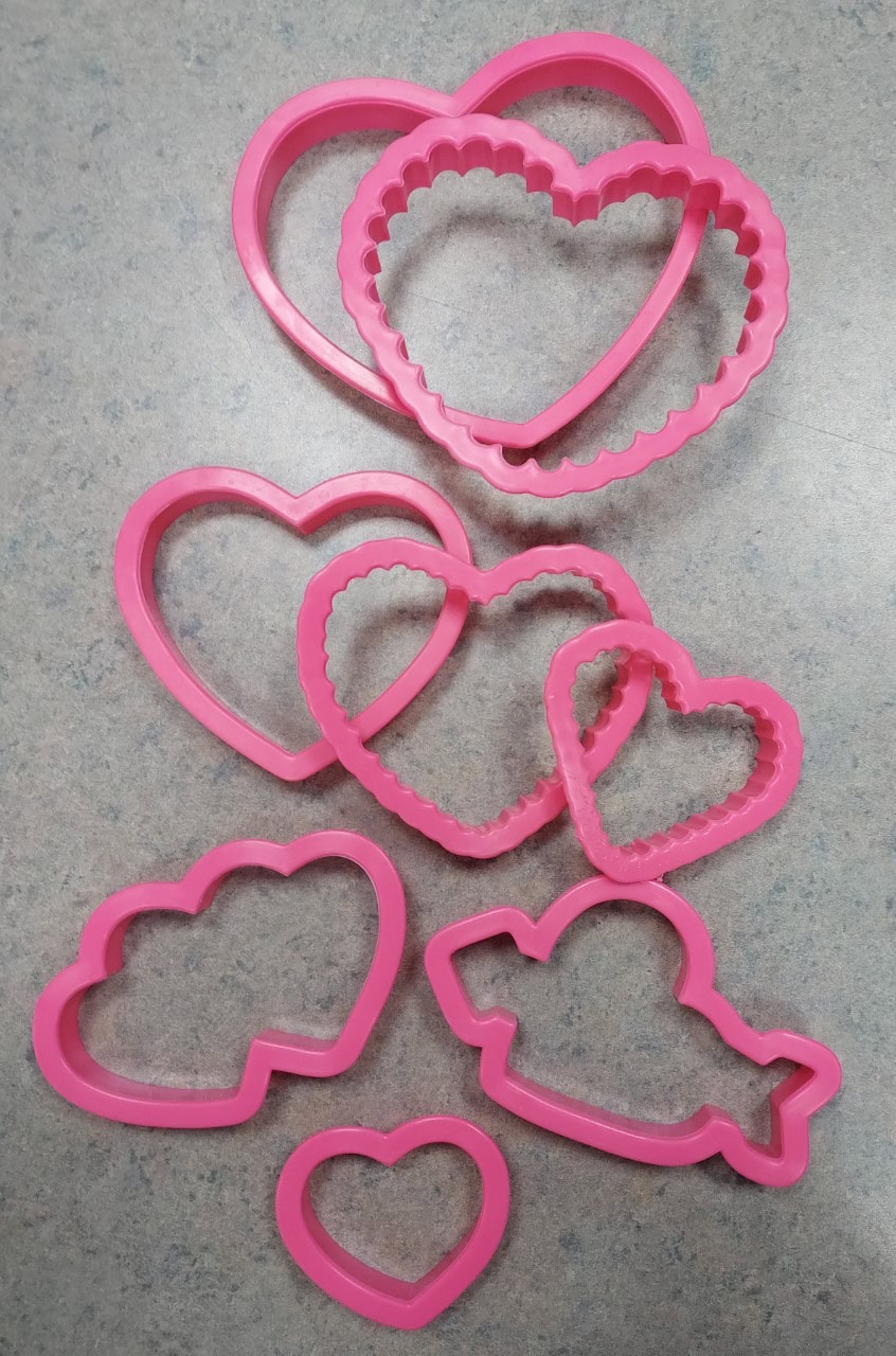 Several pink plastic cookie cutters in heart, cloud, and bird shapes are arranged on a gray speckled surface. The heart cutters come in various sizes and styles, some with scalloped edges.
