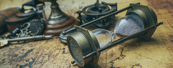A vintage hourglass with sand partially fallen, lying on an old map surrounded by antique objects, including a compass, a pen, and various metal items.