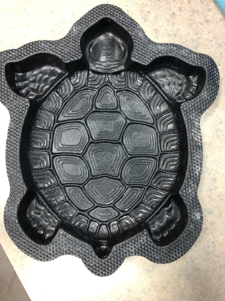 A black plastic mold in the shape of a turtle, showing its shell, legs, head, and tail, placed on a light-colored surface.