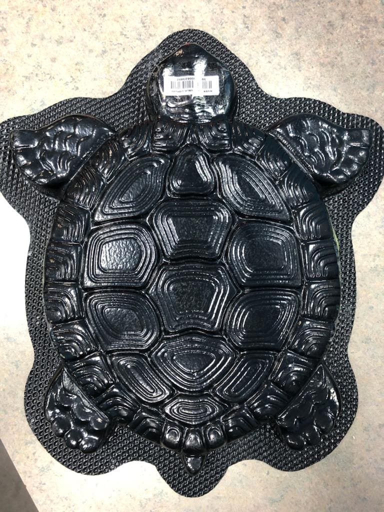 A black, turtle-shaped tray or mold with detailed shell and limb patterns lies on a gray, speckled surface. A barcode sticker is placed on the turtles head.