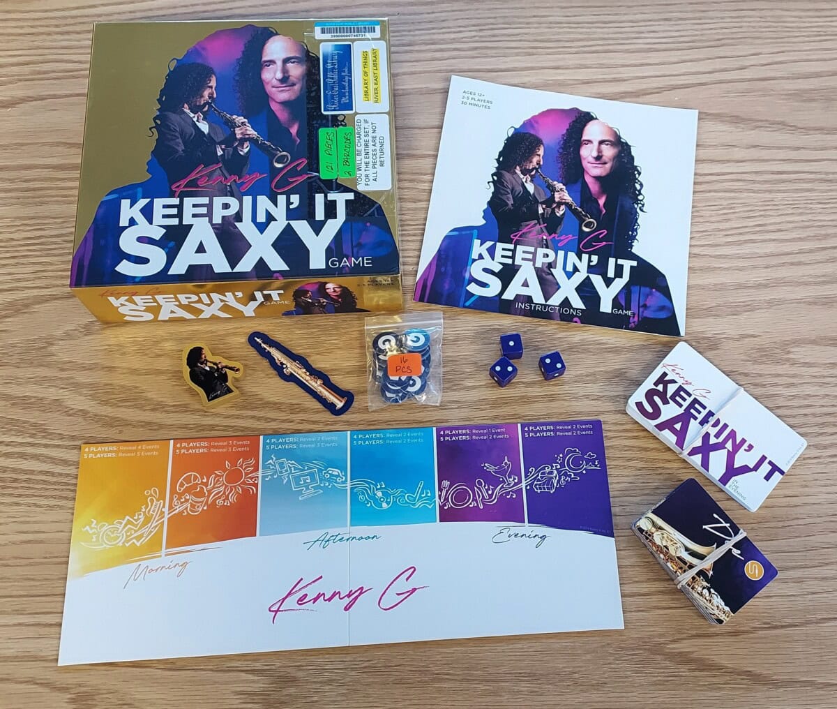 Kenny G-themed board game Keepin It Saxy on a wooden table, showing the box, instruction booklet, game pieces, dice, cards, and illustrated player boards labeled Morning, Afternoon, and Evening.