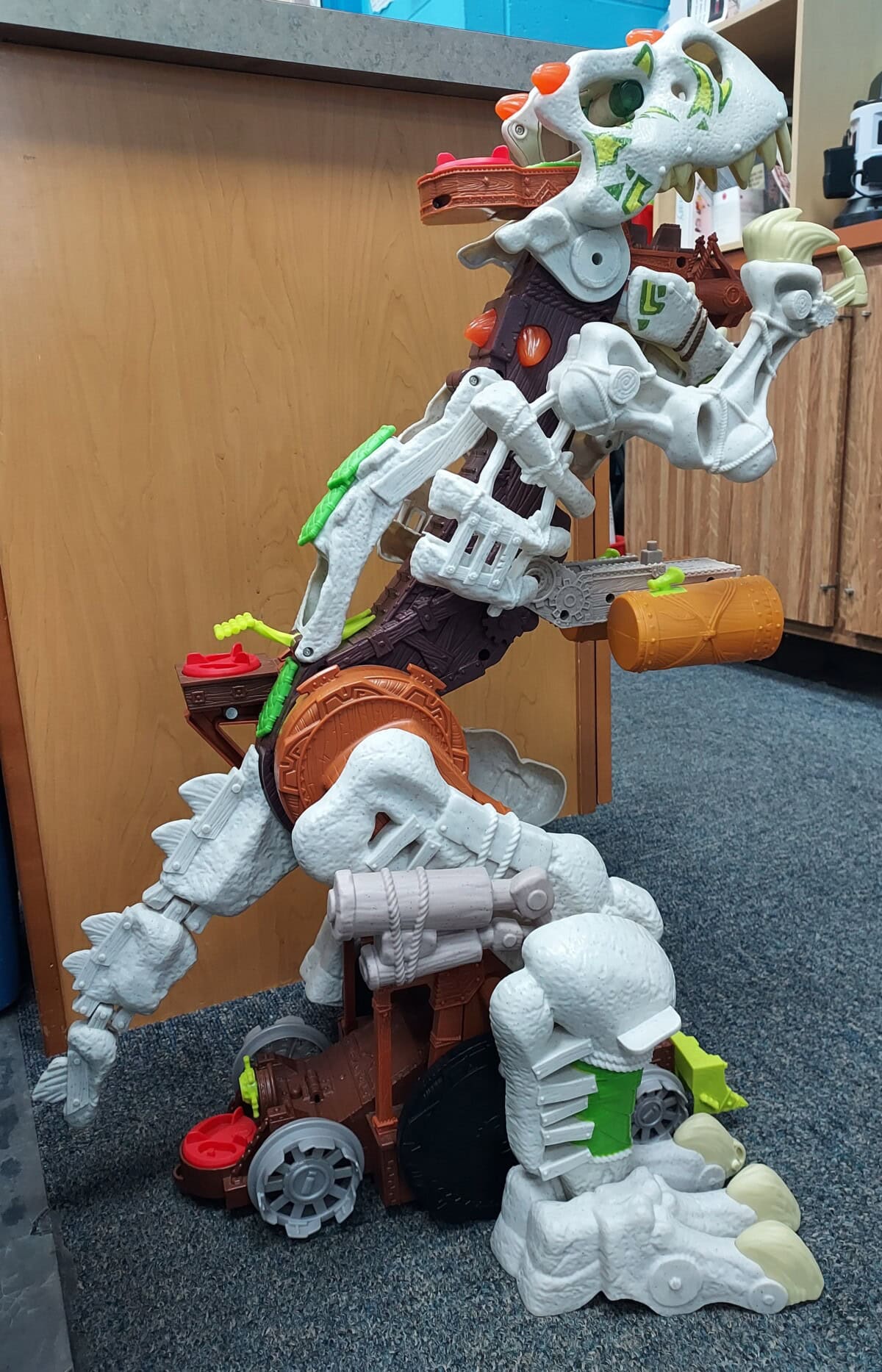 A large toy dinosaur made of white, brown, and green plastic parts, featuring wheels, orange and green accents, and mechanical details, is positioned upright next to a wooden cabinet on a carpeted floor.
