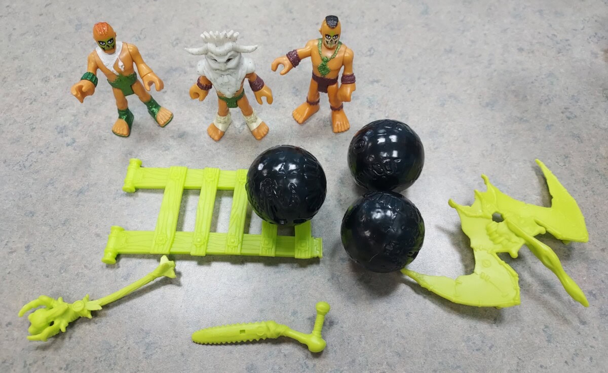 Three action figures, a green ladder, three black spheres, a green dragon, and two green plastic weapons are arranged on a gray tabletop.