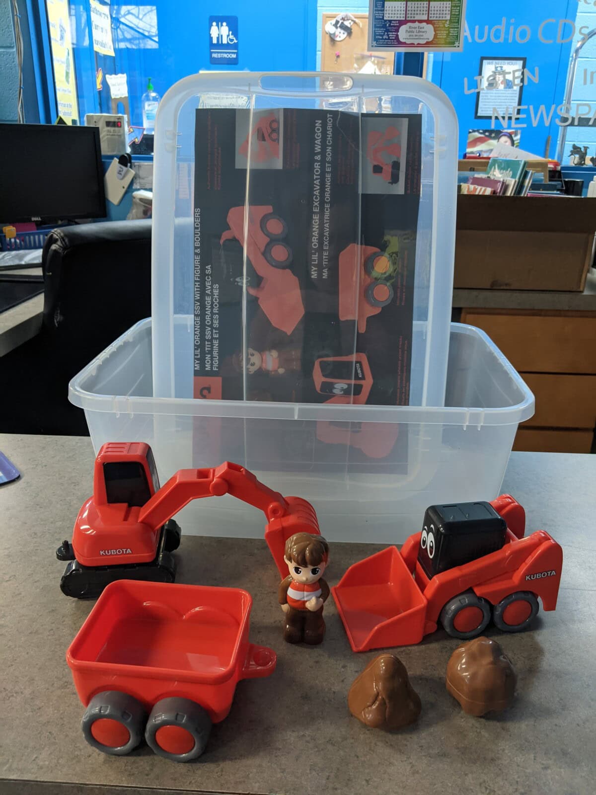 A toy construction set with a red excavator, a red front loader with eyes, a red wagon, a small brown-haired figure, and two brown rock shapes on a table. A clear plastic storage bin is in the background.