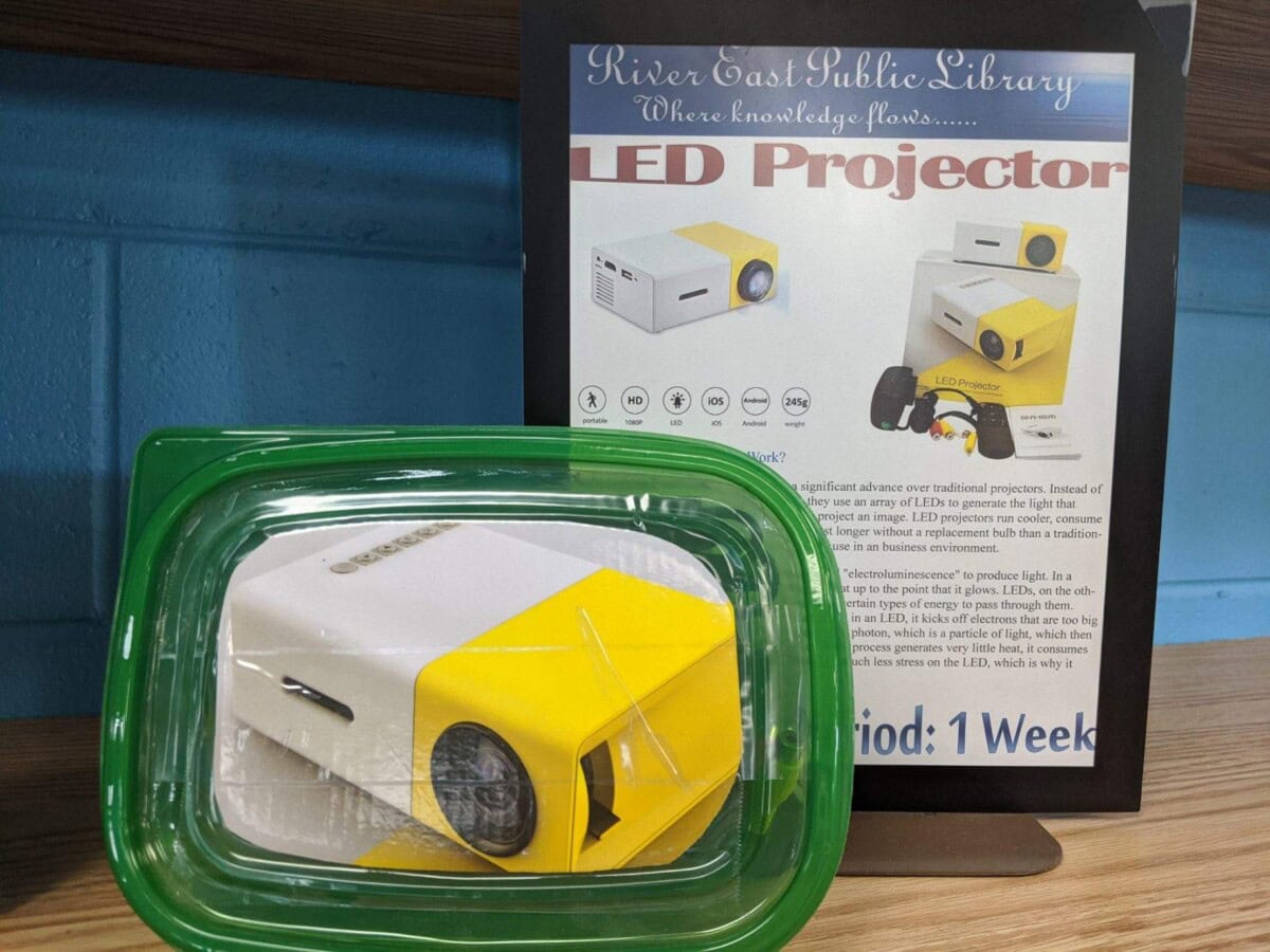 A small white and yellow LED projector in a clear plastic container, placed in front of a library sign advertising the projector. The sign features images and information about the LED projector.