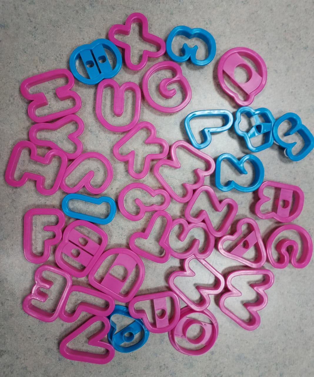 Plastic cookie cutters shaped like blue numbers and pink letters are scattered on a gray surface. The numbers and letters are outlined, and some overlap each other.