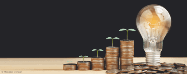 Stacks of coins increasing in height, each topped with a small plant sprout, are arranged left to right. A lit lightbulb sits beside the tallest stack, symbolizing growth and innovation.
