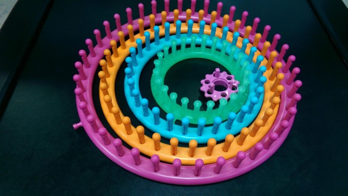 Four round plastic knitting looms of different sizes and colors (pink, orange, blue, and green) are stacked inside each other on a black background. Each loom has evenly spaced pegs around the outer edge.