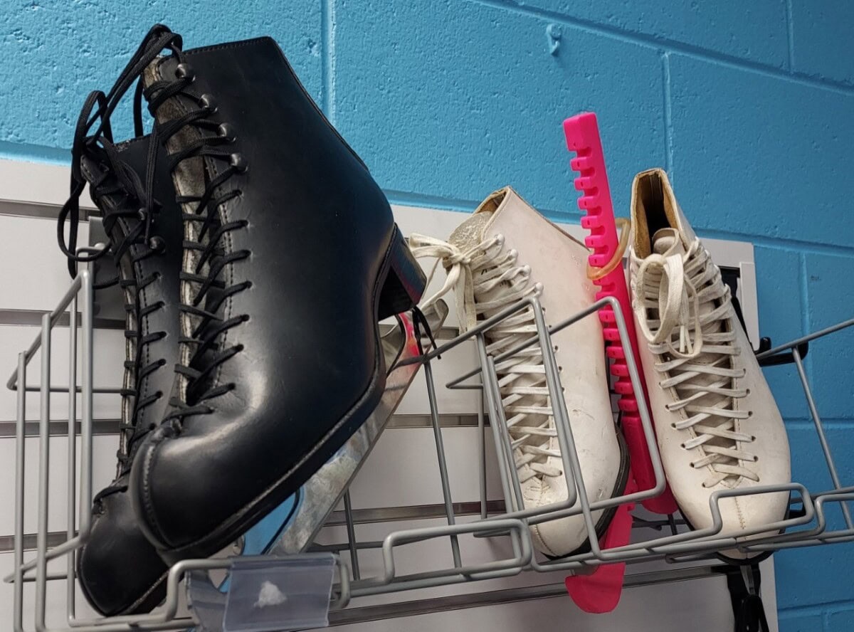 A pair of black ice skates and a pair of white ice skates with pink blade guards rest on a metal rack against a blue wall.