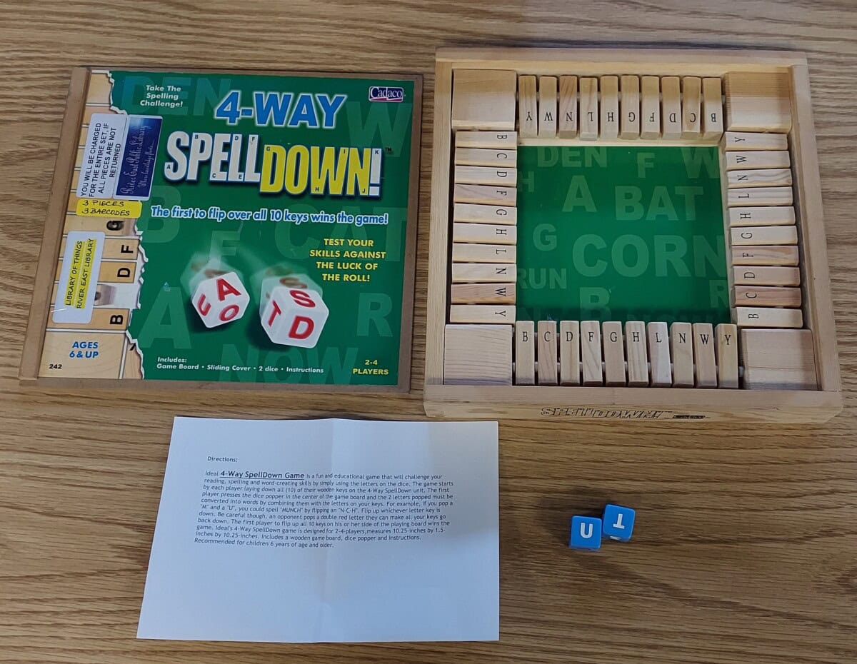 A board game called 4-Way SpellDown! is shown open on a wooden table, with wooden letter tiles in a tray, letter dice, the game box, and instructions placed nearby.