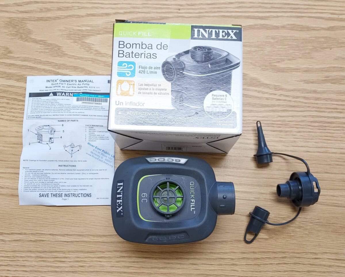 Intex Quick-Fill battery-powered air pump with nozzle attachments, owners manual, and product box displayed on a wooden surface. The packaging and manual include text in English and Spanish.