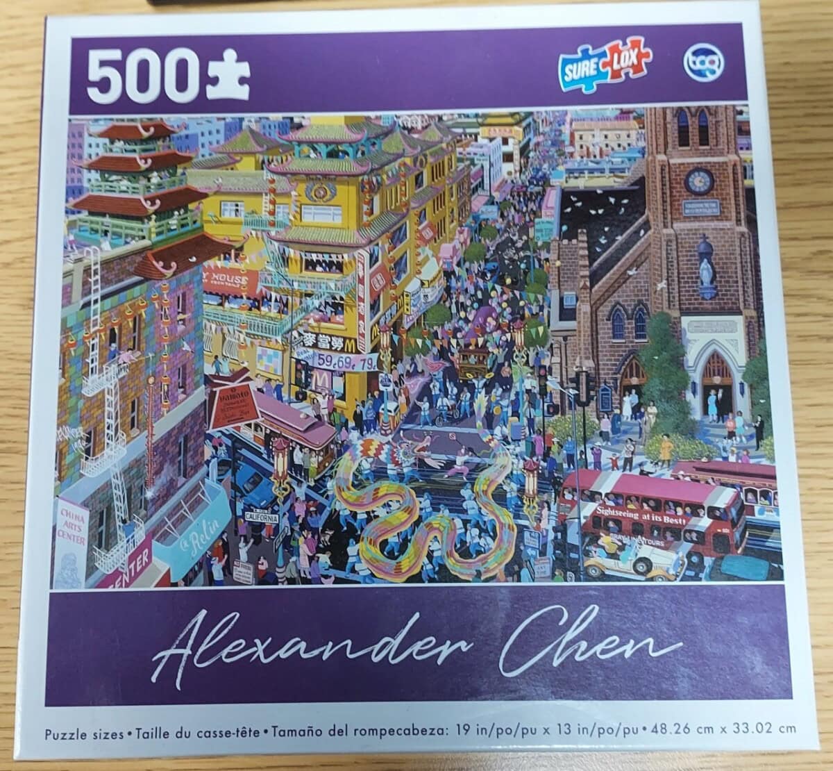 A 500-piece puzzle box featuring a colorful Alexander Chen illustration of a bustling city street with a parade, dragon dancers, people, shops, and vehicles, including a double-decker bus, under a purple sky.