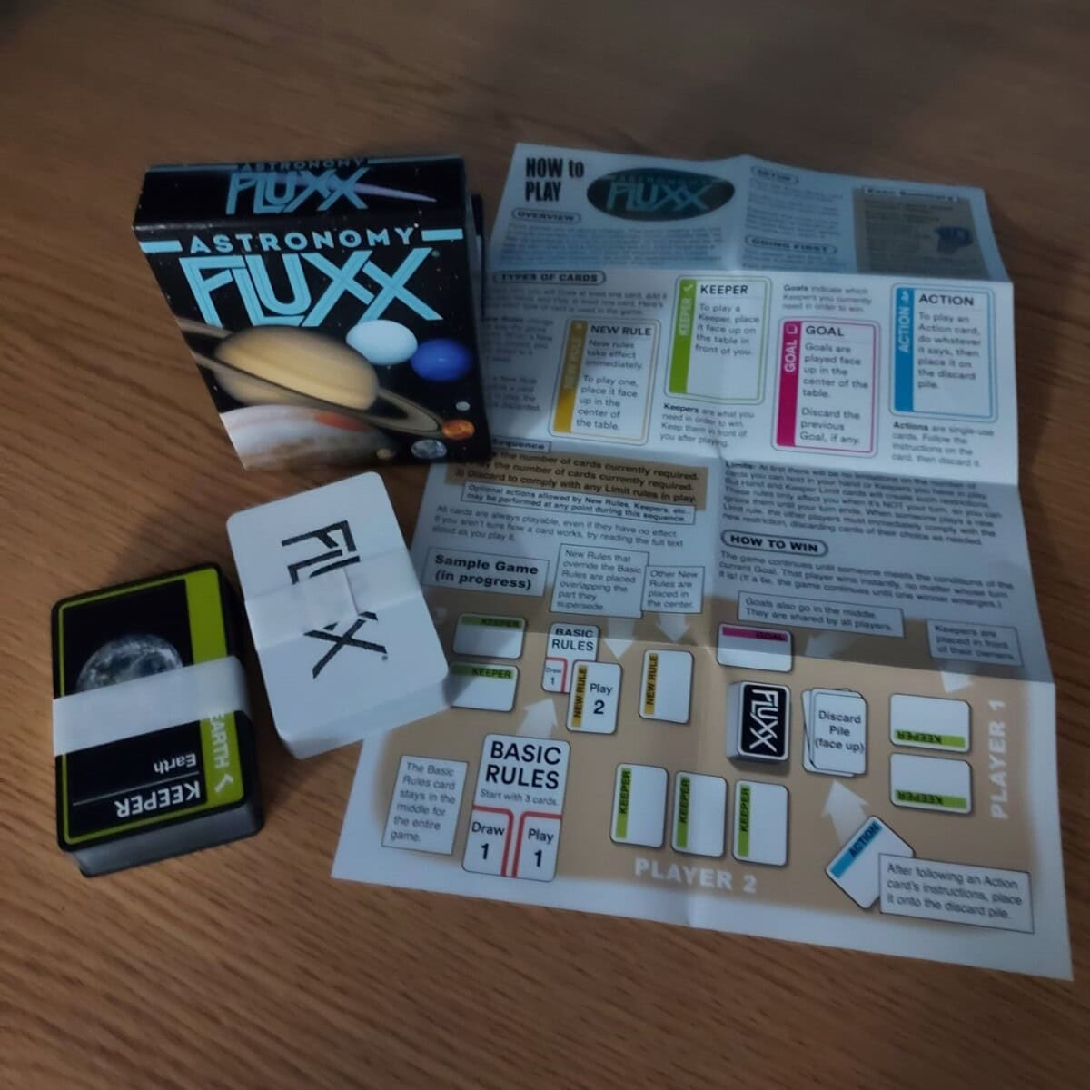 A game of Astronomy Fluxx is set up on a wooden table with the card deck, box, rule sheet, and cards labeled “FLUXX” and “KEEPER” visible, along with a game board showing rules and actions.