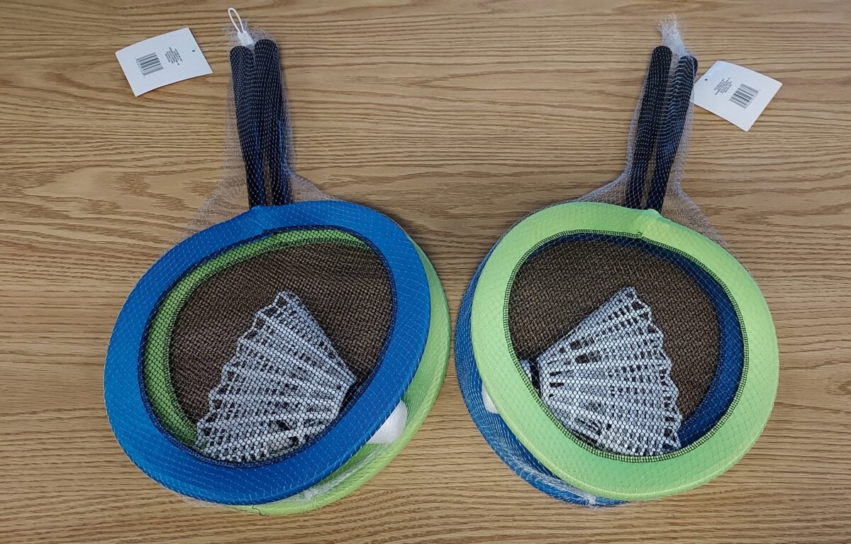 Two sets of badminton rackets, each with a birdie and colorful rings (one blue and one green), are wrapped in netting and placed on a wood surface. Each set has a white tag attached to the handle.