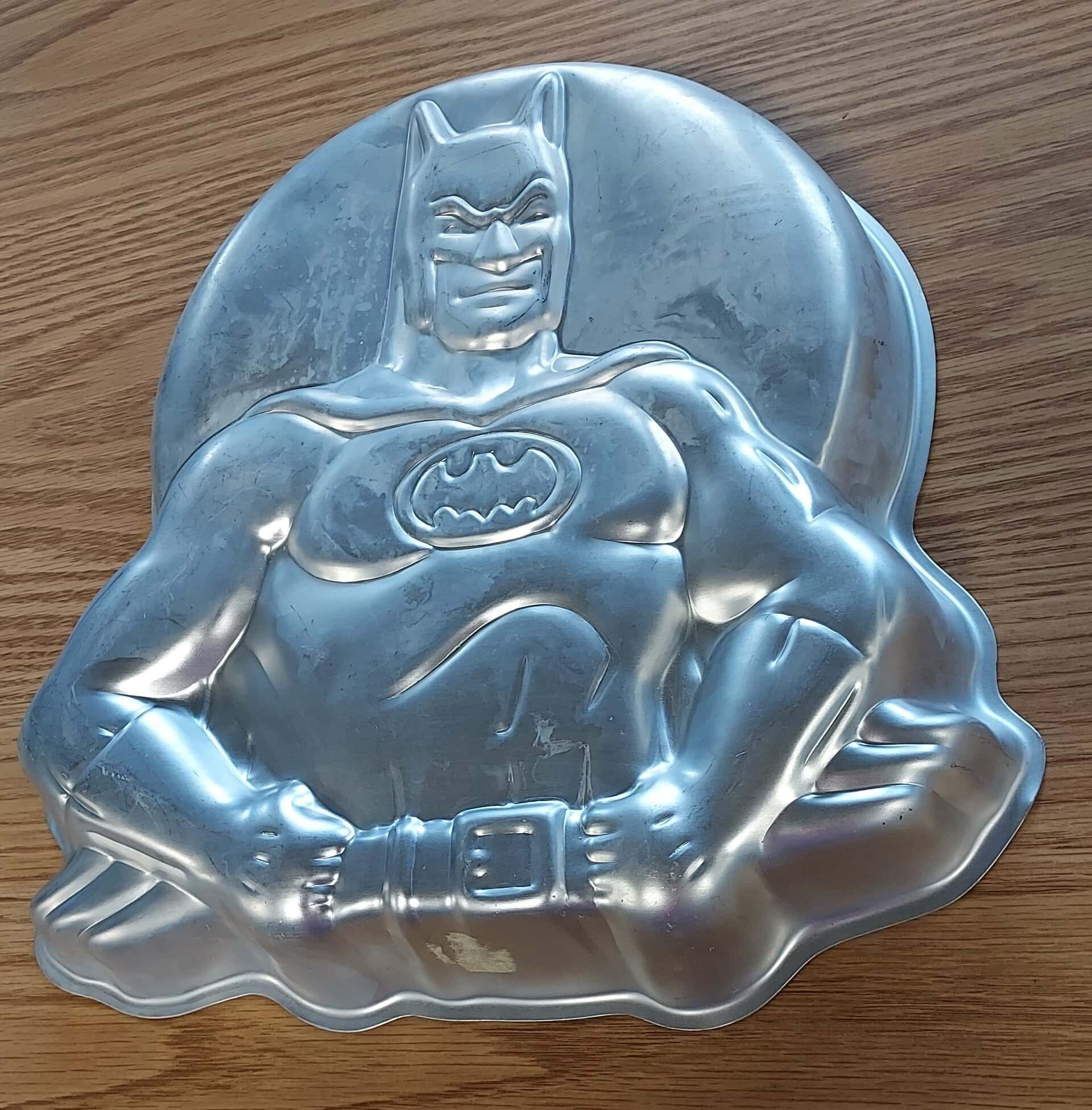 A metal cake pan shaped like Batman, showing a muscular Batman figure with the bat symbol on his chest, lying on a wooden surface.