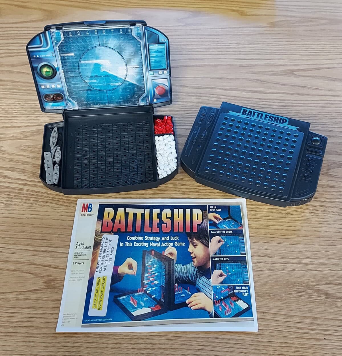A Battleship board game set on a wooden table, showing two open game boards with red and white pegs, and a game instruction sheet with an image of children playing.