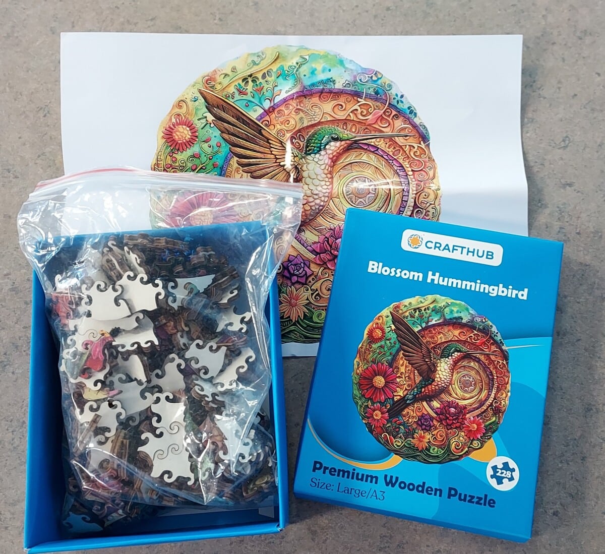 A box of CraftHubs Blossom Hummingbird wooden puzzle pieces, its blue packaging, and a reference image showing a colorful hummingbird surrounded by flowers. The puzzle is labeled for ages 12 and up.