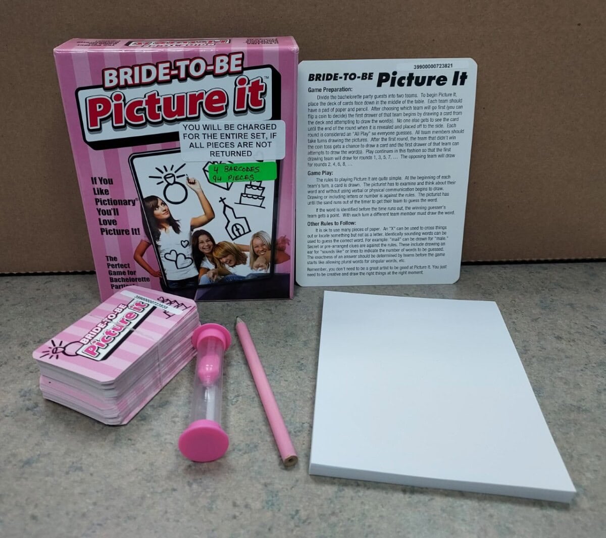 A Bride-to-Be Picture It party game set, including a pink box, instruction card, stack of cards, pink pencil, pink sand timer, and blank notepad, arranged on a gray surface against a cardboard background.