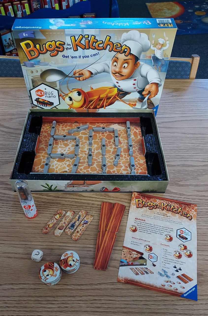 A Bugs in the Kitchen board game set on a table, showing the game box, a maze board inside the box, game pieces, a die, tokens, a bug figurine in a tube, and an instructions sheet.