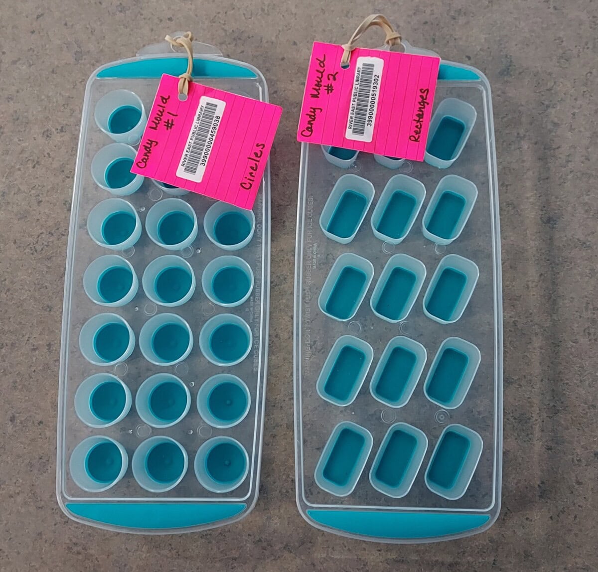 Two blue ice cube trays with different shapes—one with circles, one with rectangles—sit on a gray surface. Each tray has a pink tag labeled Candy Wheel, with additional handwritten notes attached by a ring.