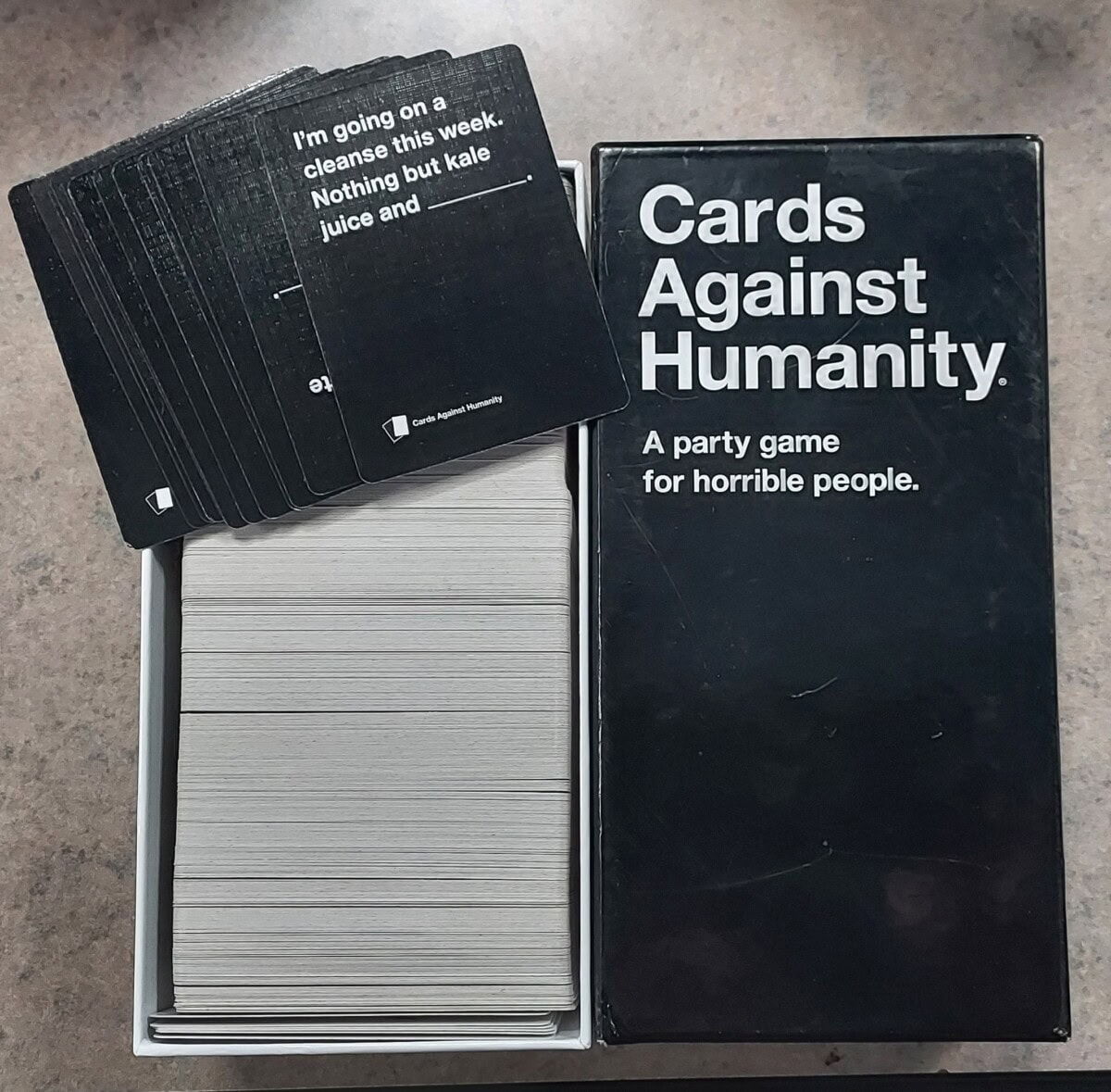 An open box of Cards Against Humanity sits on a tabletop. Several black cards are fanned out, showing prompts, while a large stack of white cards fills the box. The lid reads, Cards Against Humanity. A party game for horrible people.