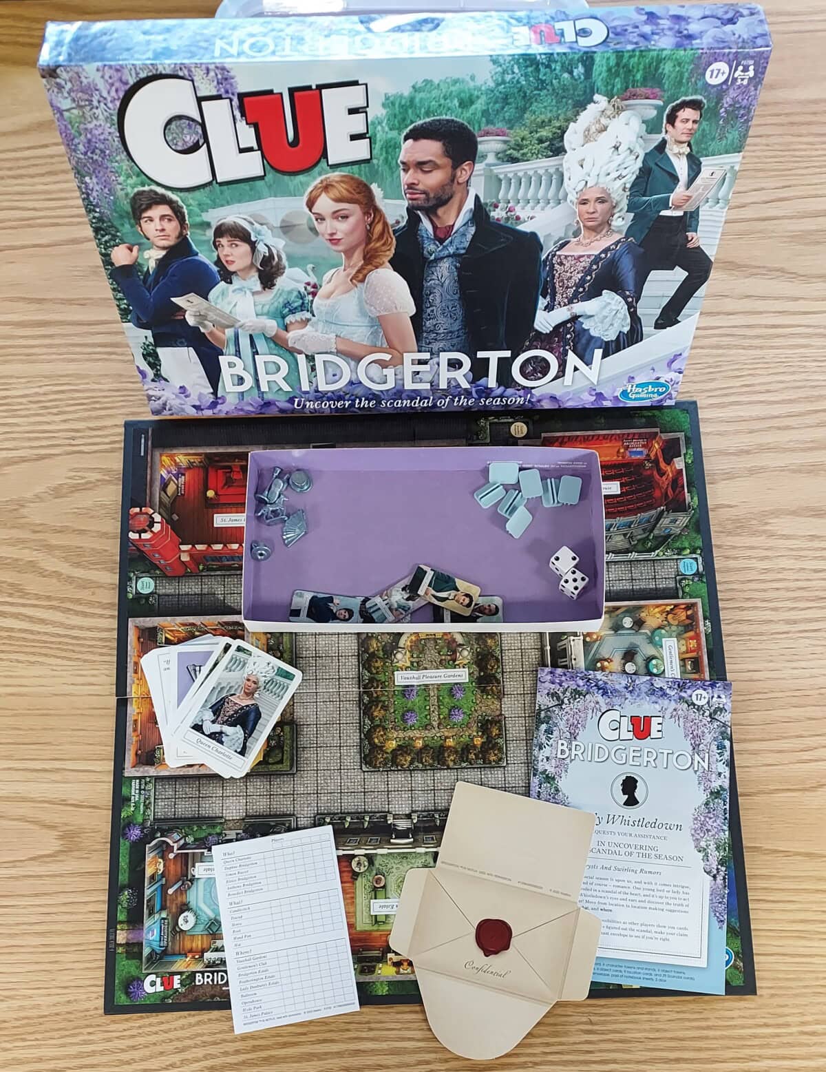 The board game Clue: Bridgerton edition is open on a wooden surface, showing the game board, character cards, playing pieces, dice, instruction sheet, a score pad, and an envelope with a red seal.