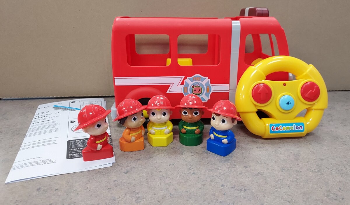 A red toy fire truck with a yellow steering wheel and five firefighter figurines in different colored outfits. An instruction sheet lies on the surface beside the toys.