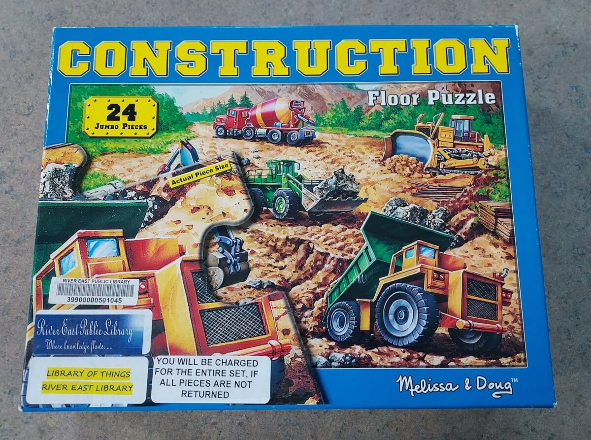 A colorful box for a 24-piece “Construction” floor puzzle by Melissa & Doug, featuring illustrations of construction vehicles. Library and return stickers are on the bottom left corner of the box.