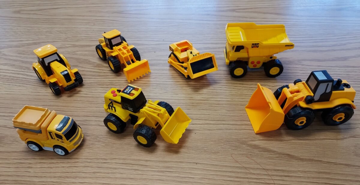 A group of seven yellow toy construction vehicles, including dump trucks, bulldozers, and loaders, are arranged on a wooden surface.