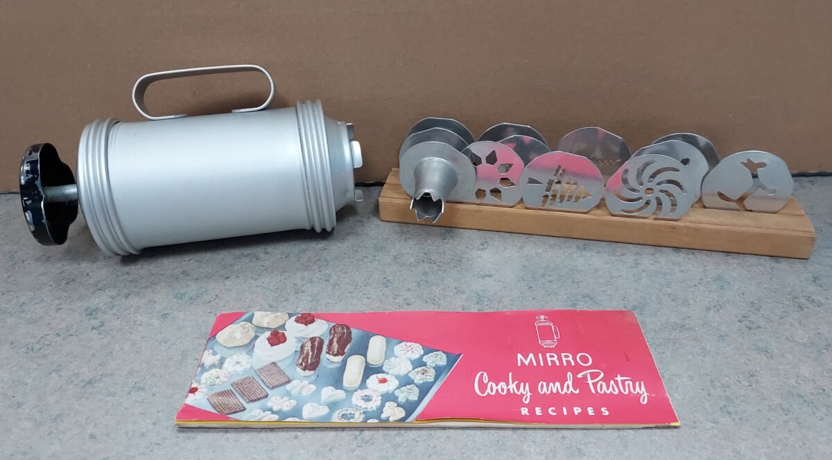 A vintage metal cookie press with various shaped discs on a wooden stand, and a pink Mirro Cooky and Pastry Recipes booklet, all placed on a gray surface with a brown background.