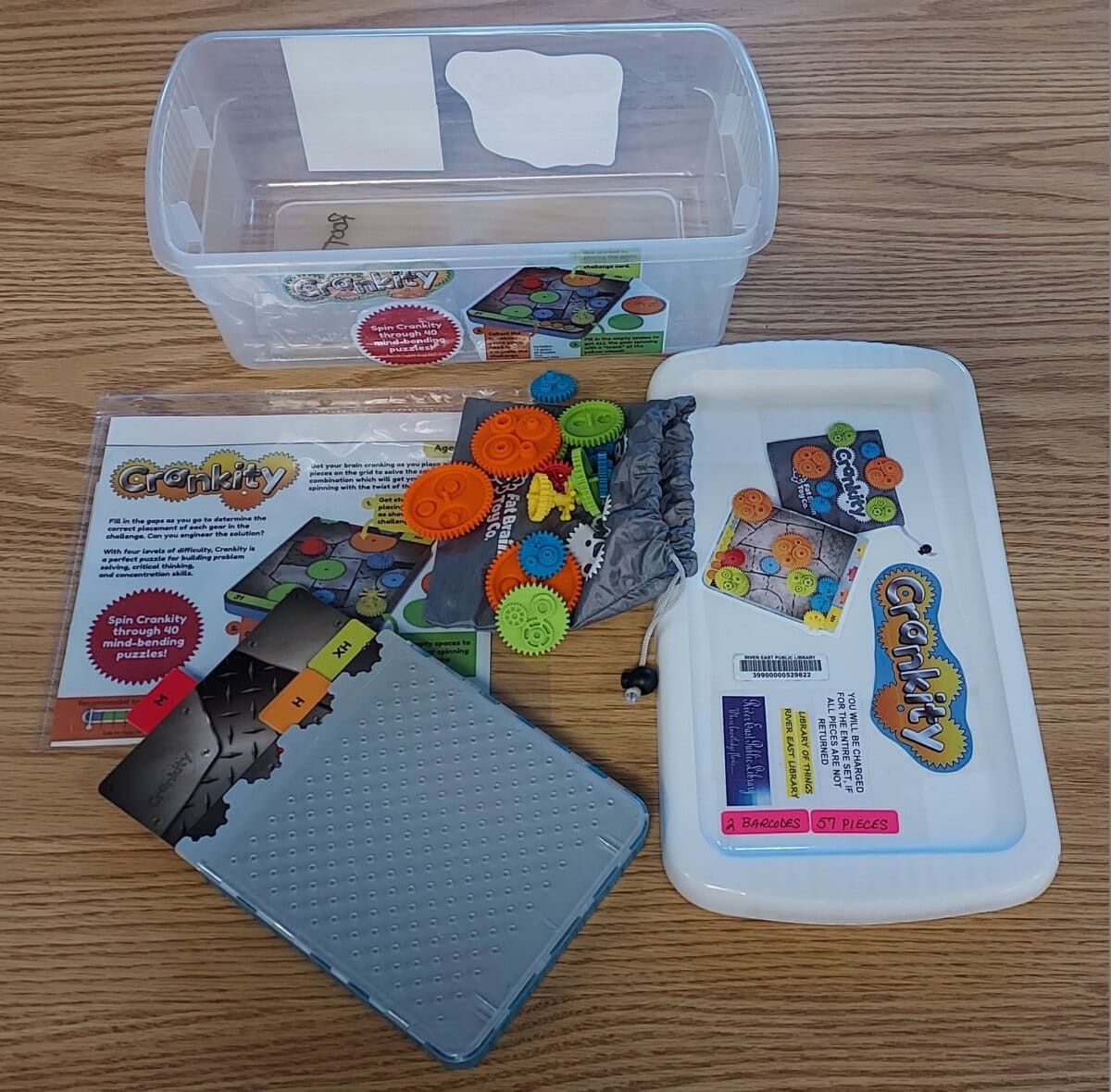 A plastic bin sits on a wooden table with a Graky STEM kit. The kit includes gears, cards, a perforated tray, instruction sheets, and colorful game pieces arranged on and around the bin lid.