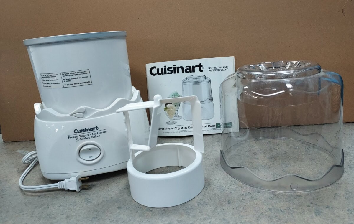 Cuisinart frozen yogurt and ice cream maker with base, paddle, clear plastic lid, and instruction manual displayed on a countertop against a brown background.