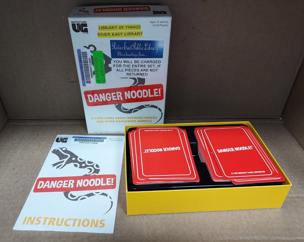 A card game set titled Danger Noodle! with a box, a stack of red cards, and an instruction sheet with a snake illustration, all placed in a cardboard container. The box has a library label and warning notice.