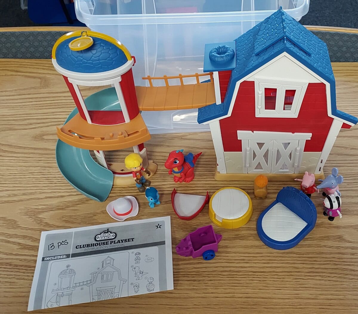 A toy clubhouse playset with a slide, bridge, barn, animal and human figures, and accessories on a wooden table. An instruction sheet labeled Clubhouse Playset is also visible.