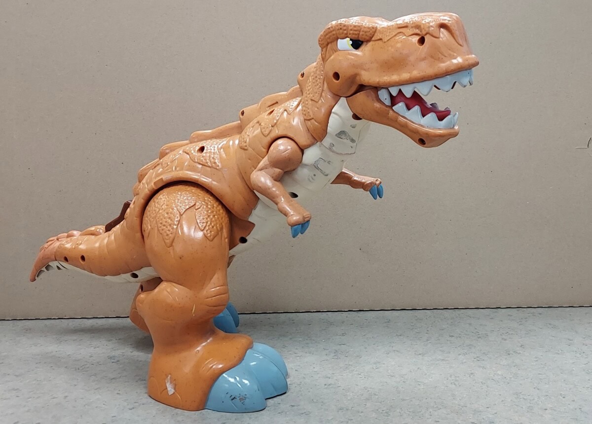 A plastic toy dinosaur, resembling a T-Rex, stands on a gray surface with its mouth open. The toy is mostly brown with some blue on its claws and a white underbelly. The background is a plain beige wall.