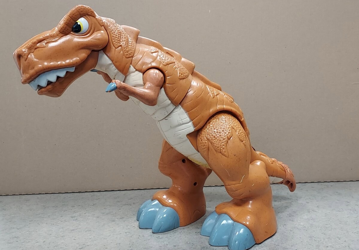 A brown and orange plastic toy dinosaur with big blue feet, a white underbelly, and a friendly expression stands on a gray surface against a plain beige background.