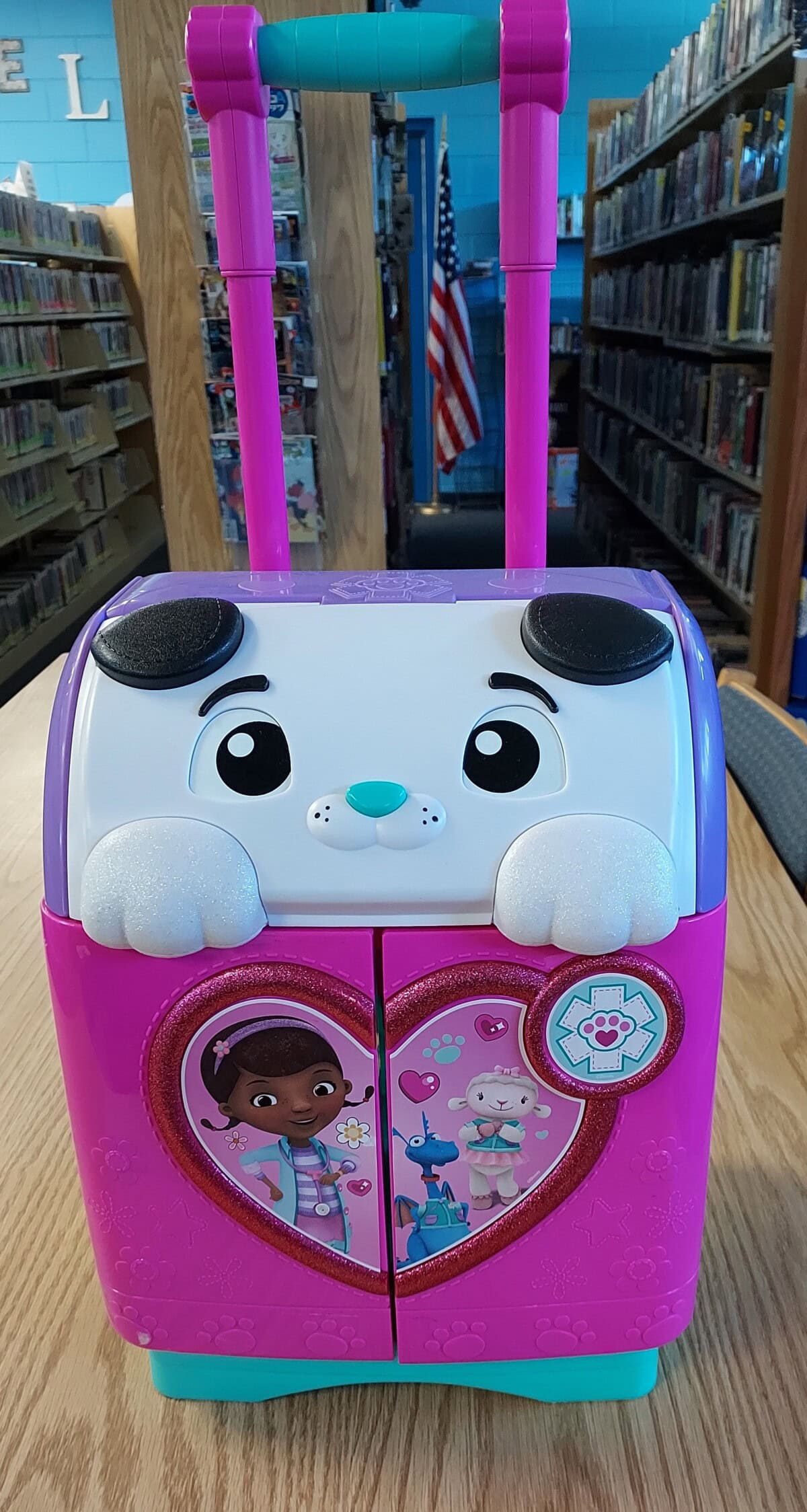 A pink childrens rolling suitcase with a cute dog face design, featuring a handle and heart-shaped decoration with Doc McStuffins and her toy friends. The suitcase is on a library table with bookshelves and an American flag in the background.