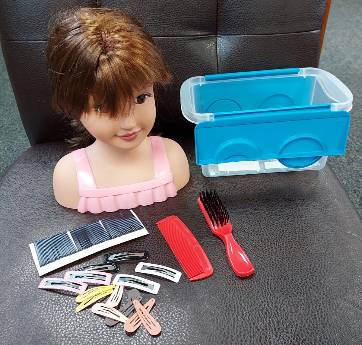 A doll head with brown hair and bangs sits on a chair next to a blue plastic container, a red comb, a black hairbrush, black hairpins, and assorted hair clips in different colors.