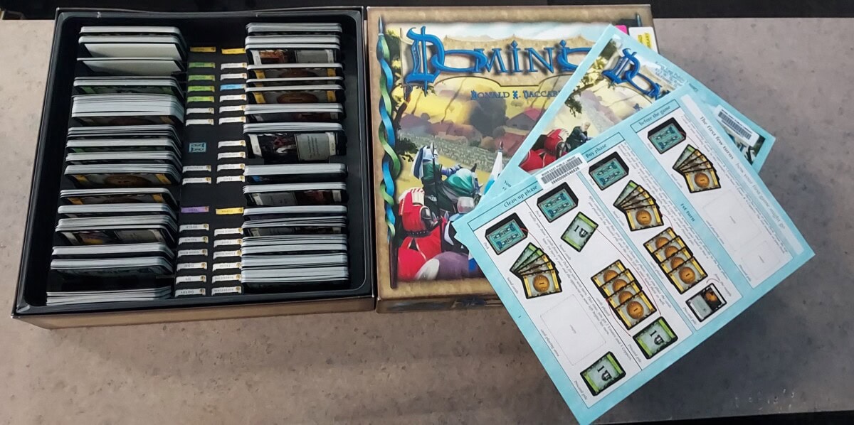 An open box of the board game Dominion is shown with stacks of cards neatly organized inside. The game box lid and several unpunched sheets of game tokens are placed on the right side of the box.