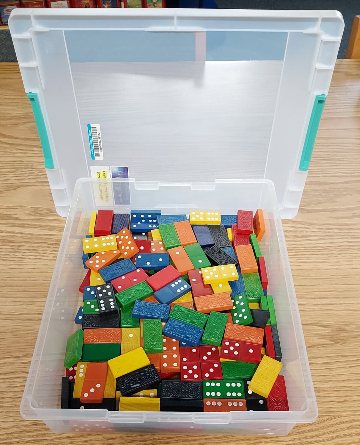 A clear plastic bin filled with colorful dominoes, including red, yellow, green, blue, and black pieces. The bin is open and sits on a wooden surface.