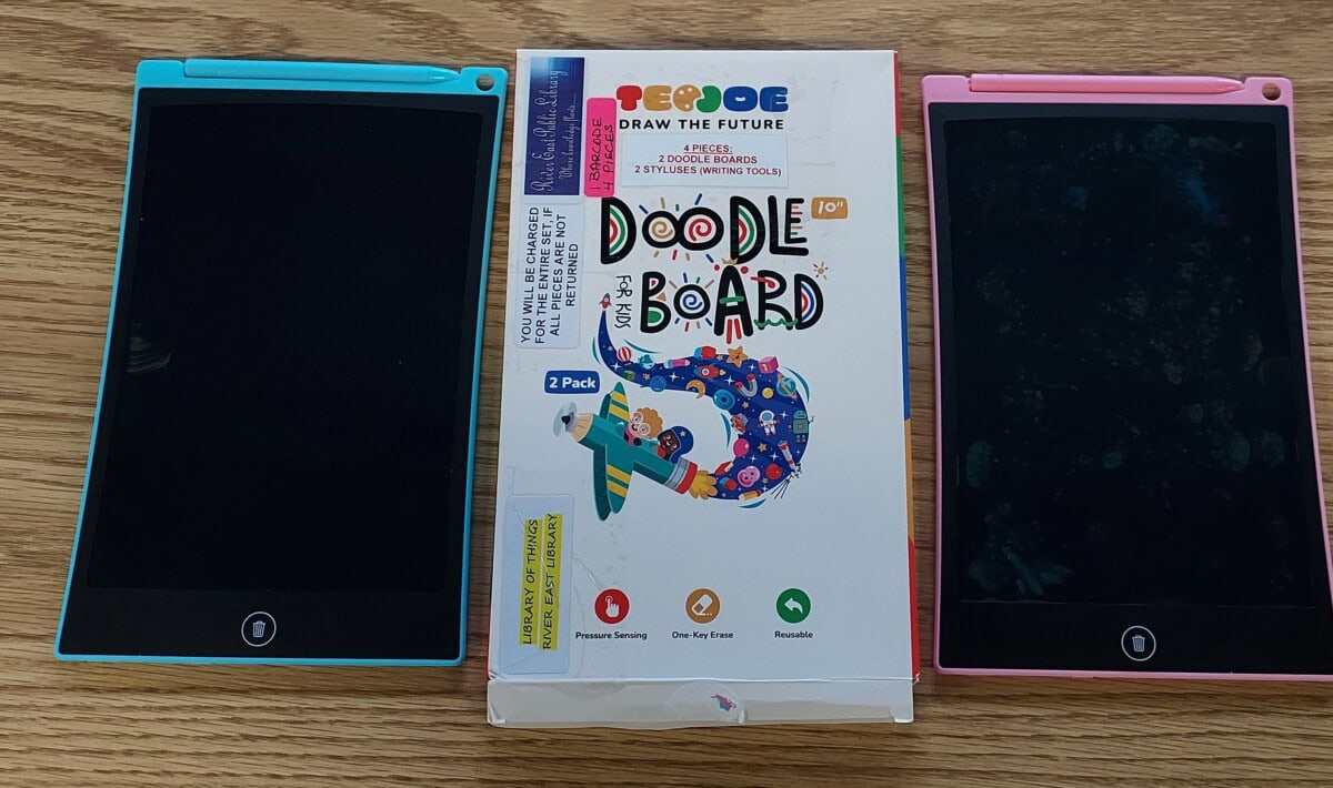 Two LCD writing tablets, one with a blue frame and one with a pink frame, are placed on either side of a colorful Doodle Board packaging box on a wooden surface.