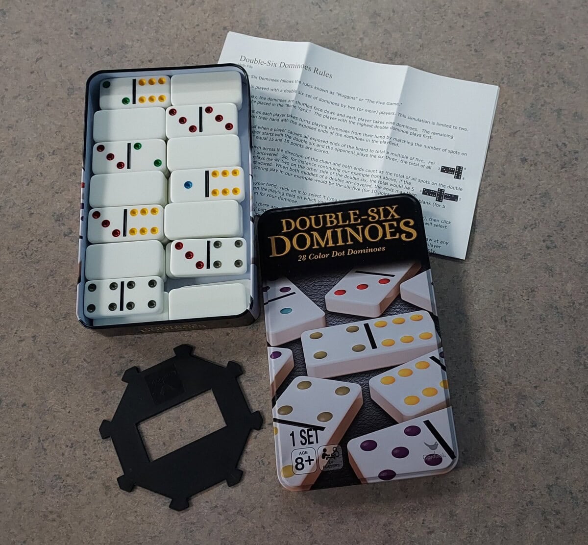 A tin of Double-Six Dominoes with colorful dots is open on a table, revealing domino tiles inside. The lid, an instruction sheet, and a black plastic domino holder are placed beside the tin.