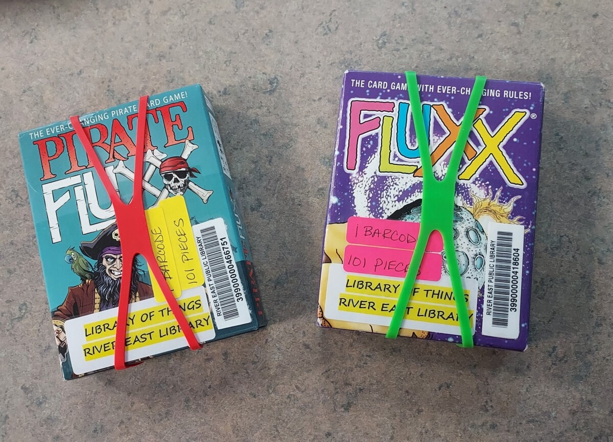Two card games, Pirate Fluxx and Fluxx, sit on a gray surface. Each is held together with rubber bands and labeled with library barcodes and tags indicating pieces and their origin from River East Library.