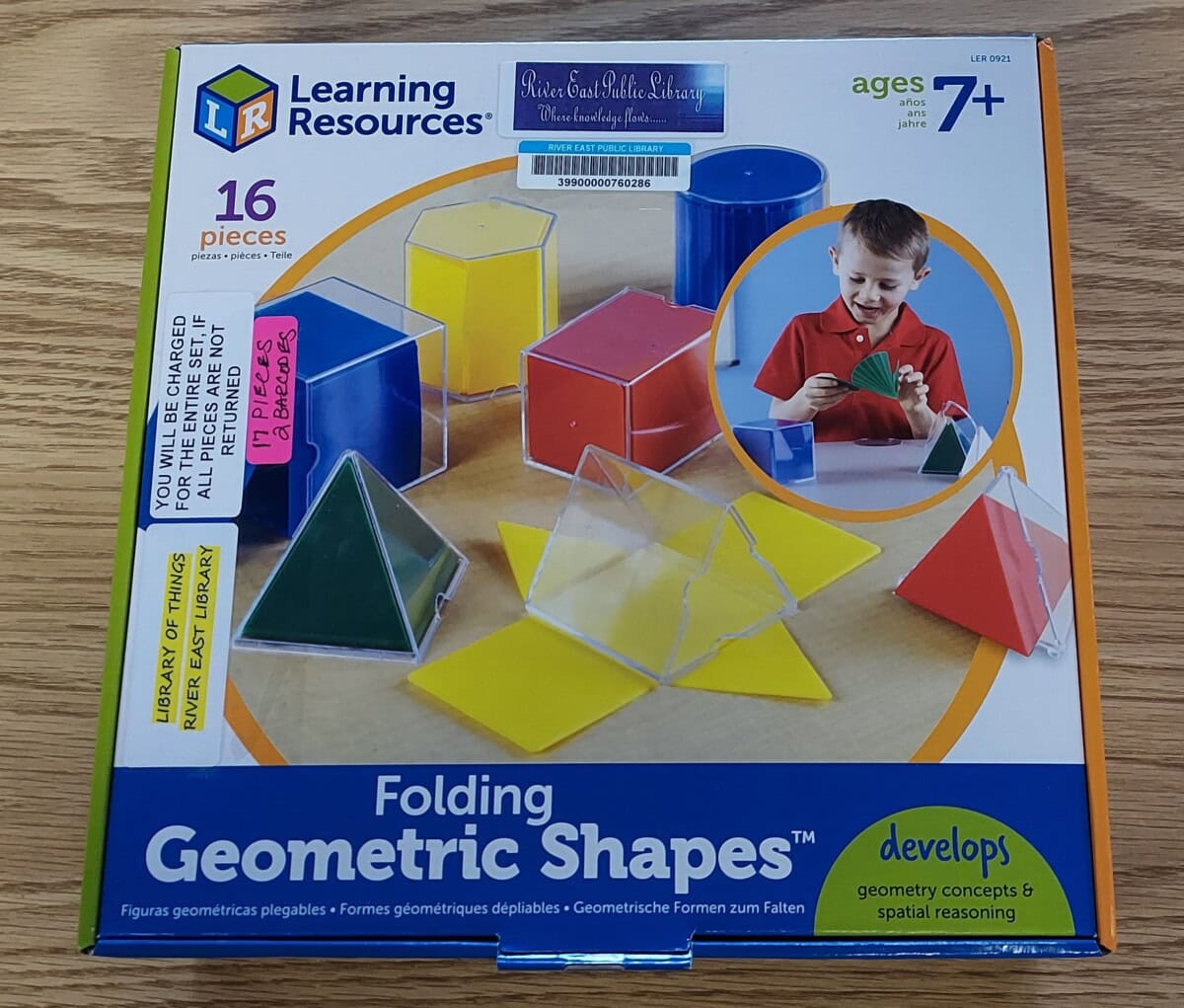 A box of Learning Resources Folding Geometric Shapes for ages 7+, showing 16 pieces, various colorful geometric solids, and a child assembling a shape. The box has library stickers and shelf labels on the front.