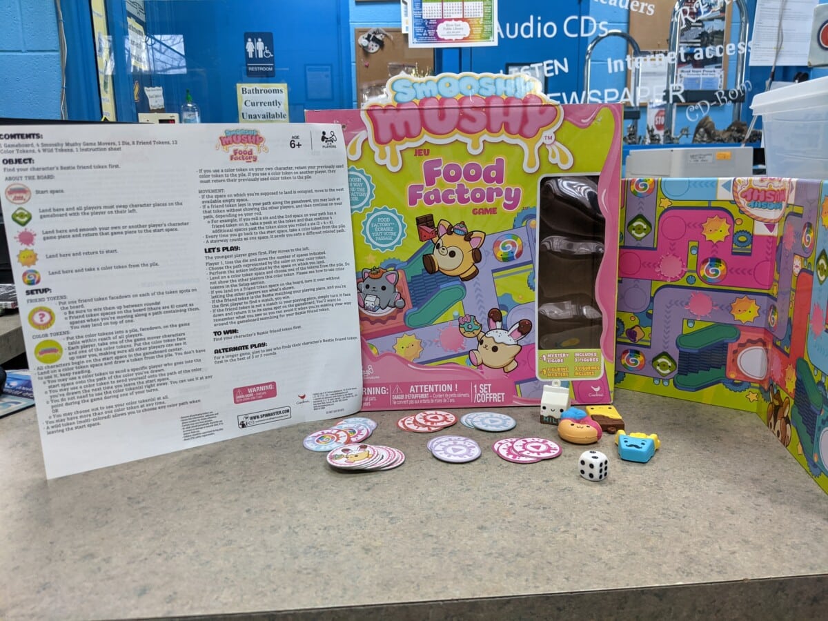 The Smooshy Mushy Food Factory board game is on display with its box, game board, instructions, dice, character pawns, and game tokens arranged on a countertop in front of a blue wall.