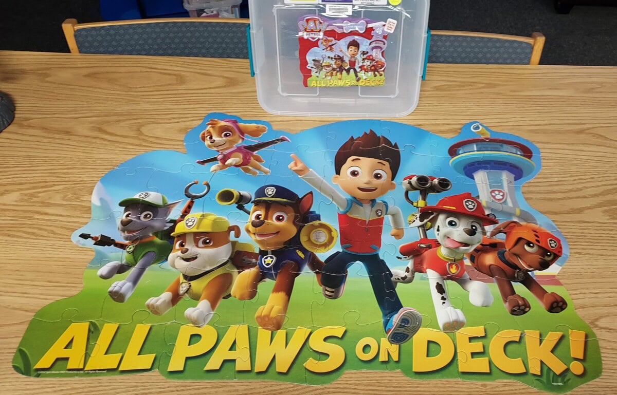 A completed Paw Patrol puzzle features six main characters and the words “ALL PAWS ON DECK!” A Paw Patrol puzzle box sits on a wooden table in the background.