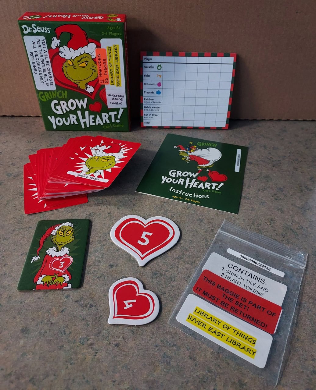 A Dr. Seuss Grinch Grow Your Heart! card game is displayed with the box, instructions, heart-shaped tokens, playing cards, a score pad, and a labeled library bag.