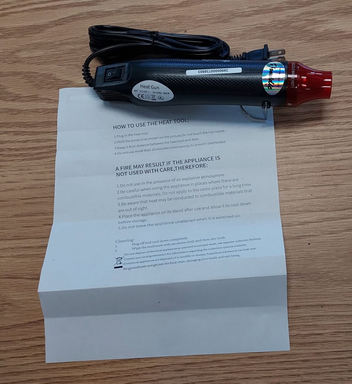 A black and red heat gun with a power cord is placed on a wooden surface, resting above a sheet of instructions on how to use the appliance safely and cleaning guidelines.