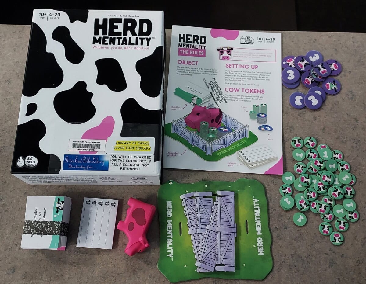 Game components for Herd Mentality displayed on a table, including the box, rule booklet, pink cow figure, cards, cow-pattern tokens, scoring pads, and a green corral board.